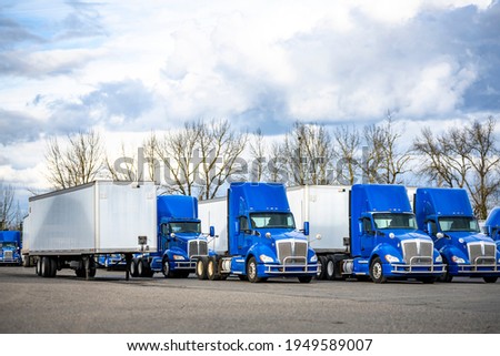 Team of professional commercial day cab blue big rig semi trucks tractors and dry van semi trailers standing at row on industrial parking lot waiting for next freight route Royalty-Free Stock Photo #1949589007