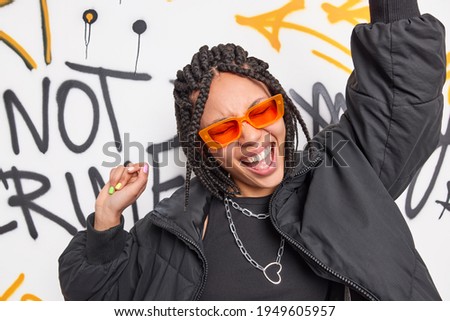 Hipster teen girl dances to pop music raises arms has fun sings song wears trendy orange sunglasses black jacket shows some movements against graffiti wall. Urban culture and modern generation Royalty-Free Stock Photo #1949605957
