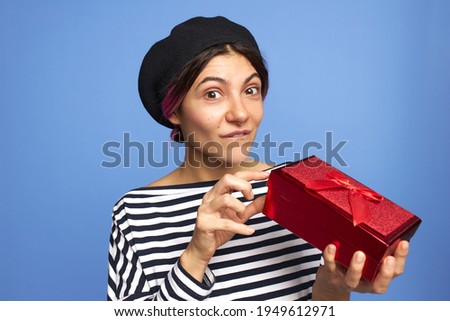 Studio image of cute happy young female with pinkish hair and nose ring raising eyebrows in amazement holding red box getting present from secret admirer, unpacking gift having curious surprised look