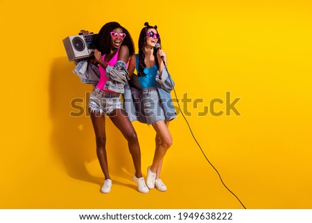 Photo of two cute ladies wear jeans outfit dark eyewear listening boom box singing looking empty space isolated yellow color background