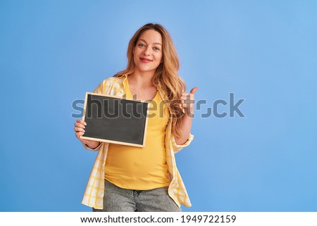 young, attractive, pregnant woman holds a board with inscriptions in her hands. Happy mom-to-be holds an empty ad banner, and points a thumbs up at the blue studio background, copy space