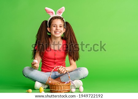 charming smiling Caucasian teenage girl sits on a green background in the studio and holds a basket of Easter eggs. A happy pre-teen child wears rabbit ears on Easter Day. easter egg hunt. Copy space