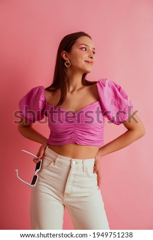 Elegant fashionable woman wearing trendy fuchsia color blouse with puff sleeves, white jeans, holding sunglasses, posing on pink background 