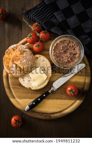 Canned liver sausage spread from long term pantry with crispy bread roll bun, butter, tomatoes and knife for snack or breakfast on cutting board and dark wooden background