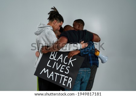 Rear view of young multiracial people hugging each other while standing on light background and holding anti racist quotes placards in hands. Stop racism concept