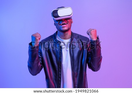 Overjoyed Black Millennial Man In Virtual Reality VR Glasses And Leather Jacket Playing Video Games, Emotionally Reacting To Win, Standing In Neon Lighting Over Purple Studio Background, Copy Space