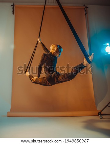 young woman in black costume is making aerial trick in longitudinal twine on the beige background in photo studio in warm and cold lights, sport concept, free space	

