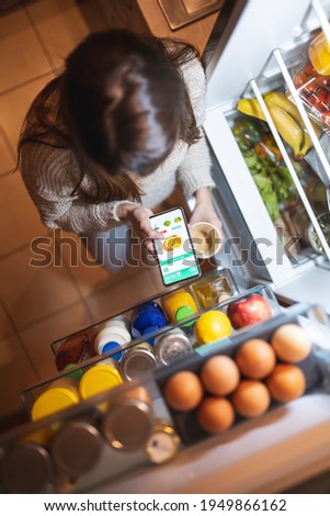 High angle view of a beautiful young woman standing next to an opened refrigerator door, ordering fresh fruit and vegetables online for home delivery using a smart phone app