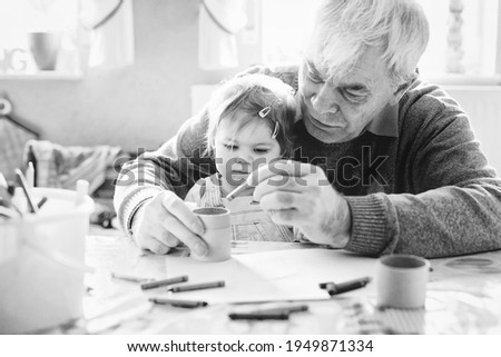 Cute little baby toddler girl and handsome senior grandfather painting with pencils at home. Grandchild and man having fun together. Family and generation in love. Old black and white image