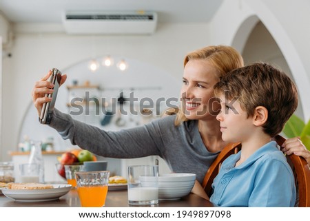 Beautiful young caucasian mother smiling and taking selfie photo using smartphone with cute little son at dining table during breakfast