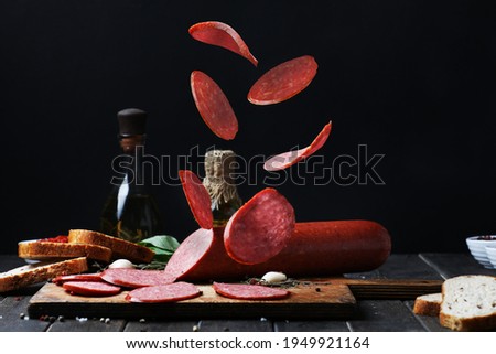 Flying Smoked Sliced Salami Sausage With Pices Of Bread,  Spices, Garlic, Oil, Vinegar On Wooden Board Black Background. Side View. Royalty-Free Stock Photo #1949921164