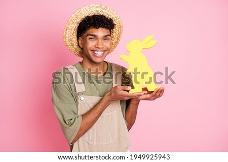 Photo portrait of curly boy in hat showing easter rabbit figure celebrating holiday smiling isolated on pastel pink color background