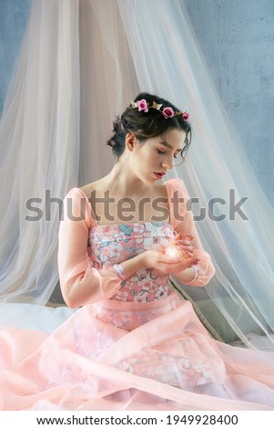 A tender young girl in the image of a princess in a pink dress with a wreath on her head holds a bright light near her heart.