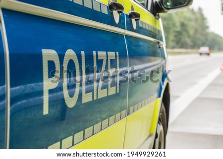 side view of a German police car on the autobahn. Lettering with reflective stripes on the body on a blue and yellow background. Rainy lanes and trees. Vehicle in the background on the hard shoulder