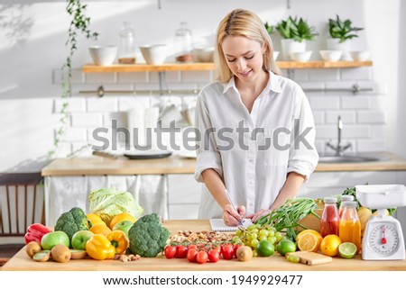 Nutritionist blonde woman writing diet plan for herself, table full of fruits and vegetables