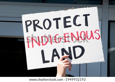 Caucasian hand holding sign reading "Protect Indigenous Land" at a protest in response to fossil fuel pipelines being routed through protected Native American reservations and danger of polluted water Royalty-Free Stock Photo #1949932192