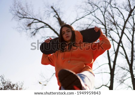 A cute teenage girl in an orange hoodie is posing with a skateboard.