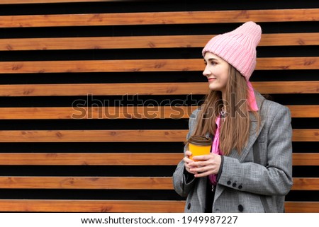 A young girl of European appearance at the age of 23 drinks coffee from a plastic disposable cup of yellow color on the street against the background of a wall of horizontal planks of orange color. 