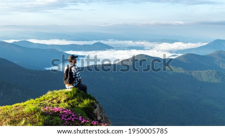 A tourist sits on the edge of a cliff covered with a pink carpet of rhododendron flowers in the summer. Foggy mountains on the background. Landscape photography