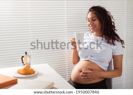 Joyful pregnant young beautiful woman looks through blinds during her morning breakfast with coffee and croissants. Concept of good morning and waiting for meeting with a baby. Copyspace
