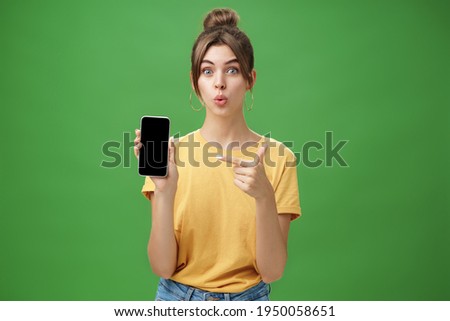 Portrait of intrigued woman cannot wait use new smartphone holding phone and pointing at cellphone screen folding lips with excitement and interest presenting new device model in store over green wall