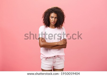 Portrait of offended gloomy sad african american female friend crossing arms on chest in protection gesture frowning looking from under forehead with insult looking envious and angry over pink wall Royalty-Free Stock Photo #1950059404
