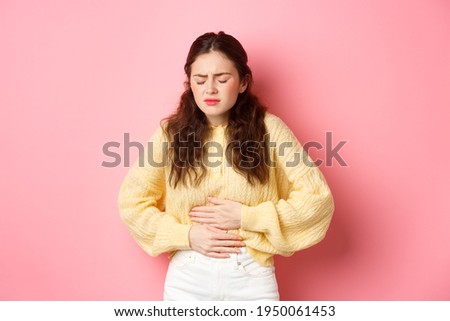 Young woman with pain in stomach, holding hands on belly, feeling terrible ache, menstrual cramps, standing against pink background Royalty-Free Stock Photo #1950061453