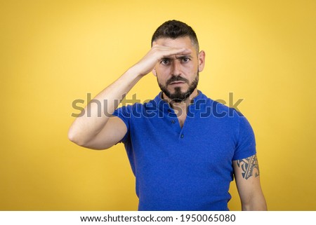 Handsome man with beard wearing blue polo shirt over yellow background very happy and smiling looking far away with hand over head. searching concept.