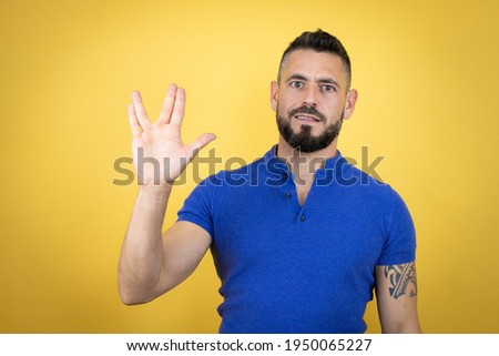 Handsome man with beard wearing blue polo shirt over yellow background doing hand symbol