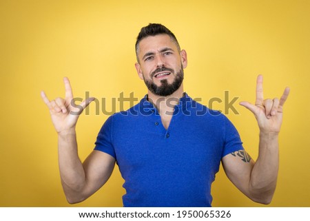 Handsome man with beard wearing blue polo shirt over yellow background shouting with crazy expression doing rock symbol with hands up