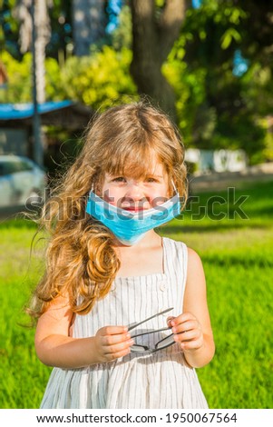 Warm summer day. Adorable little girl with long blonde curls in a protective mask. Green lawn in the park