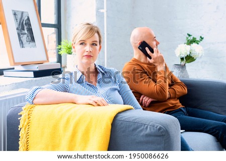 Middle age couple ignoring each other after having an argument on the sofa at home. Blond woman sitting on the couch while her husband sitting next to her and talking with somebody on his mobile phone Royalty-Free Stock Photo #1950086626