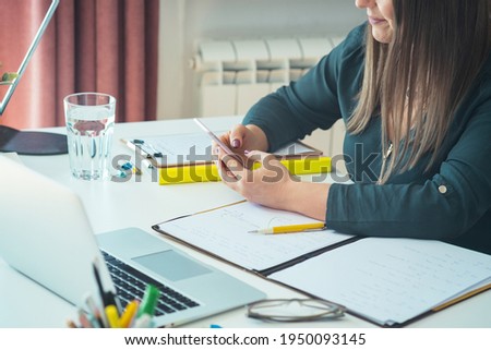 Cropped photo of unrecognizable female teacher sitting at home office desk and holding mobile phone. Young professor or student having break with online class and using smartphone to send text message