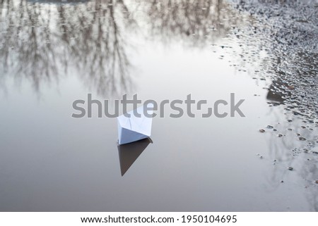 a white paper boat floats in a puddle in the reflection of the trees