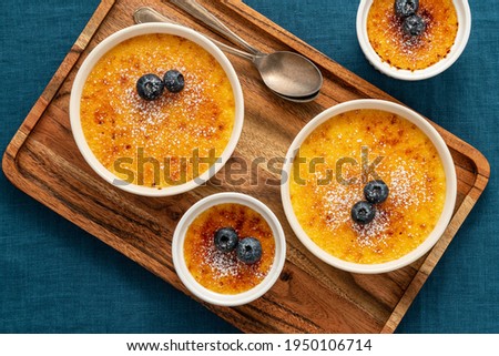 Creme brulee in lot of ramekin on wooden tray on dark linen tablecloth. Close up french vanilla cream dessert. Spanish crema catalana with layer of caramelized sugar. Top view Royalty-Free Stock Photo #1950106714