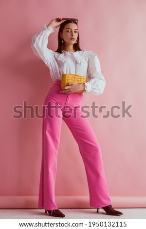 Elegant fashionable woman wearing white vintage blouse with lace collar, pink jeans, holding small padded yellow leather bag, posing on pink background. Full length portrait
 Royalty-Free Stock Photo #1950132115