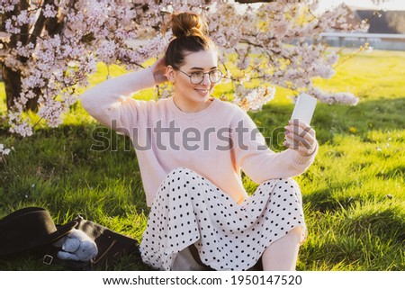 Young hipster girl in glasses making a selfie or video for a blog in social networks using mobile phone in a park near blooming tree. A laughing student having a video call during break in university
