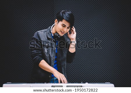 Asian male playing an electric keyboard while wearing headphones in the recording studio. Recording songs by using a studio microphone with copy space.