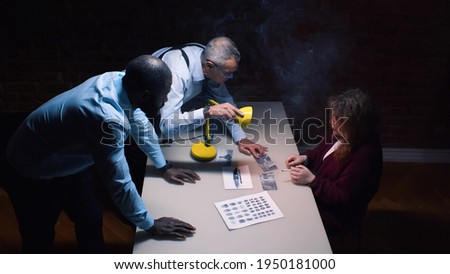 Policemen interviewing suspect woman with lamp glowing into her face. Arrested female in handcuffs being interrogated under pressure. Diverse detectives showing evidence to criminal Royalty-Free Stock Photo #1950181000