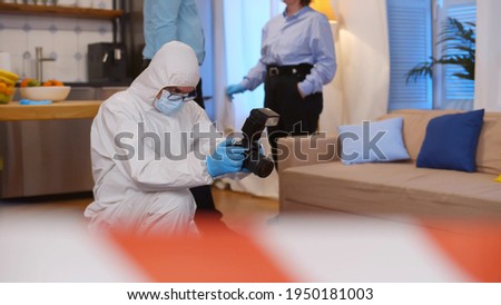 Forensic expert at crime scene doing investigation. Criminologist in safety suit taking photos while detective discussing on background working at team in modern apartment