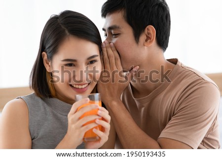 Portrait shot of cute smiling young Asian lover couple sitting on a bed together at home in the morning. Wife holding and drinking a glass of orange juice with her husband whispering her secret