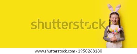 Funny happy girl with Easter bunny ears is holding colorful Easter eggs on a yellow background. Happy Easter! Copy space. Banner