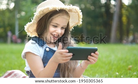 Happy smiling child girl looking in mobile phone outdoors in summer.