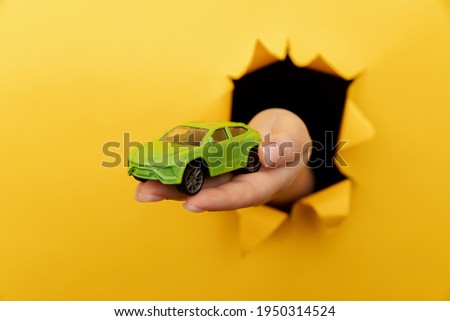 Female hand with house car through a rip in yellow paper wall close-up. Sale and rent concept