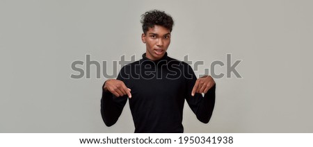 Stylish young guy dressed in black looking at camera and pointing down, presenting something, standing isolated over gray background. Advertisement concept. Copy space