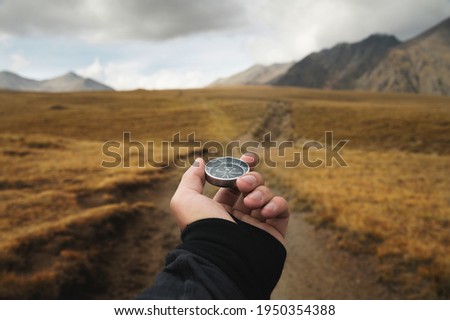 Man's hand holding a magnetic compass first-person view against the background of a high-altitude path and mountains Royalty-Free Stock Photo #1950354388