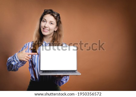 Attractive Caucasian young woman with long hair in a shirt looking at the camera holds a laptop with a cut out white screen on her hand and points her hand at the screen. Screen template blank