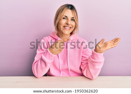 Young caucasian woman wearing casual clothes sitting on the table amazed and smiling to the camera while presenting with hand and pointing with finger. 