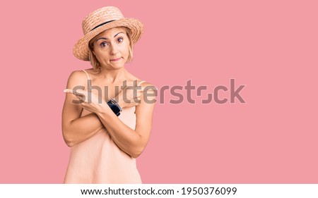 Young blonde woman wearing summer hat pointing to both sides with fingers, different direction disagree 