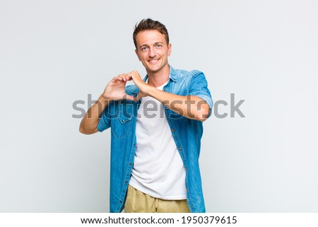 young handsome man smiling and feeling happy, cute, romantic and in love, making heart shape with both hands